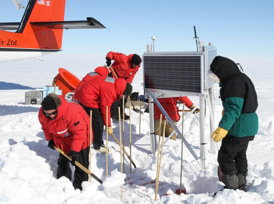 The POLENET TEAM buries a seismic station in Antarctica. The tall poles and solar array will help the next team find the station. The orange box holds batteries and the electronic equipment for the instrument. Doug Wiens, shovel in hand, is to the left. (Rachel Gesserman photo)