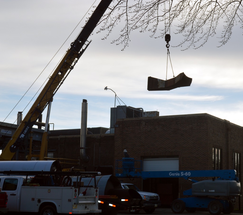 ONE OF THE old "retiring" smokestacks is lowered by crane from the roof of the Municipal Utility Power Plant. New clean energy smokestacks replaced the old. 